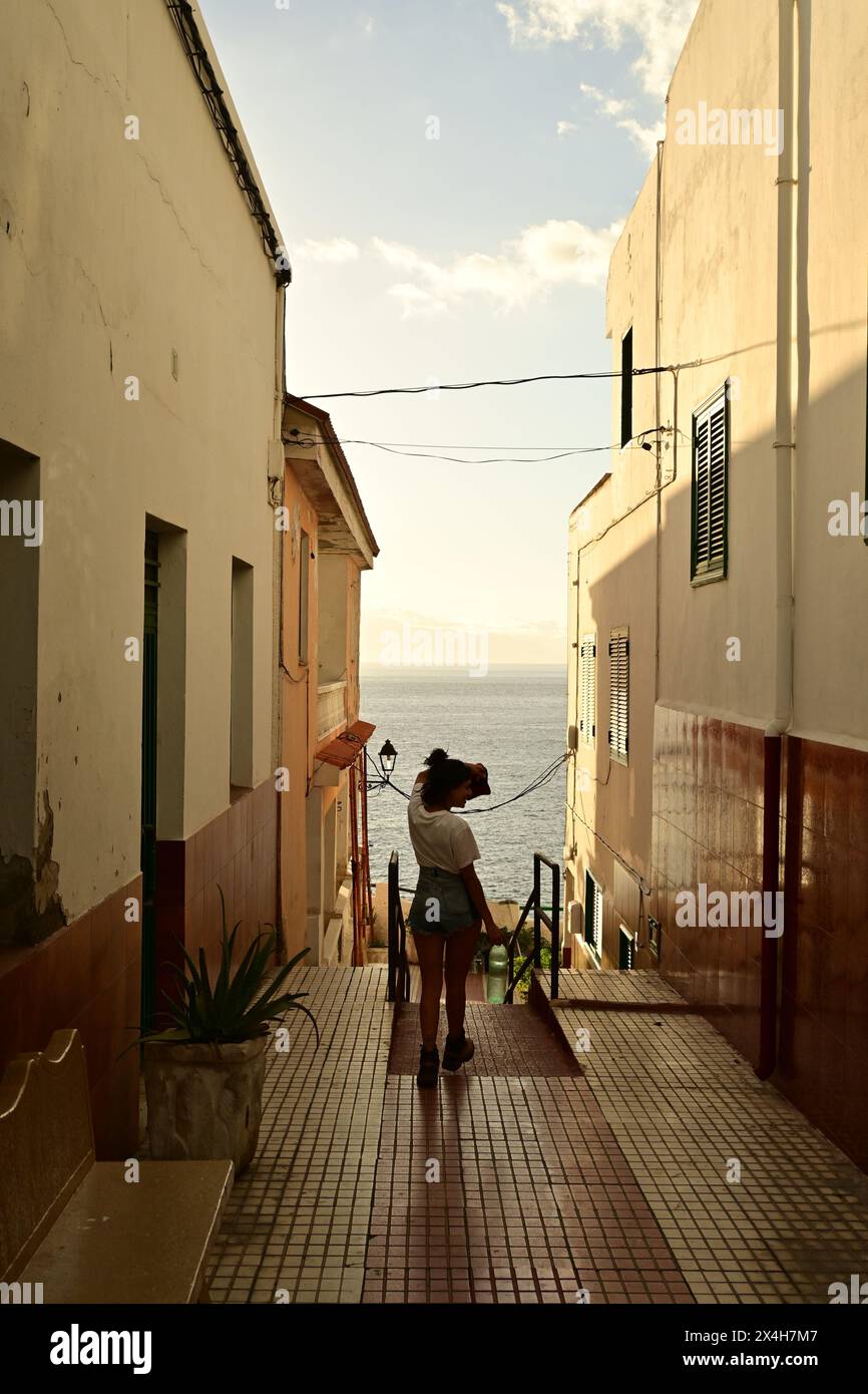 Ein Mädchen, das das Meer von einer engen Gasse aus betrachtet, genießt den Sonnenuntergang und die urbane Ruhe, umgeben von beleuchteten Wänden Stockfoto