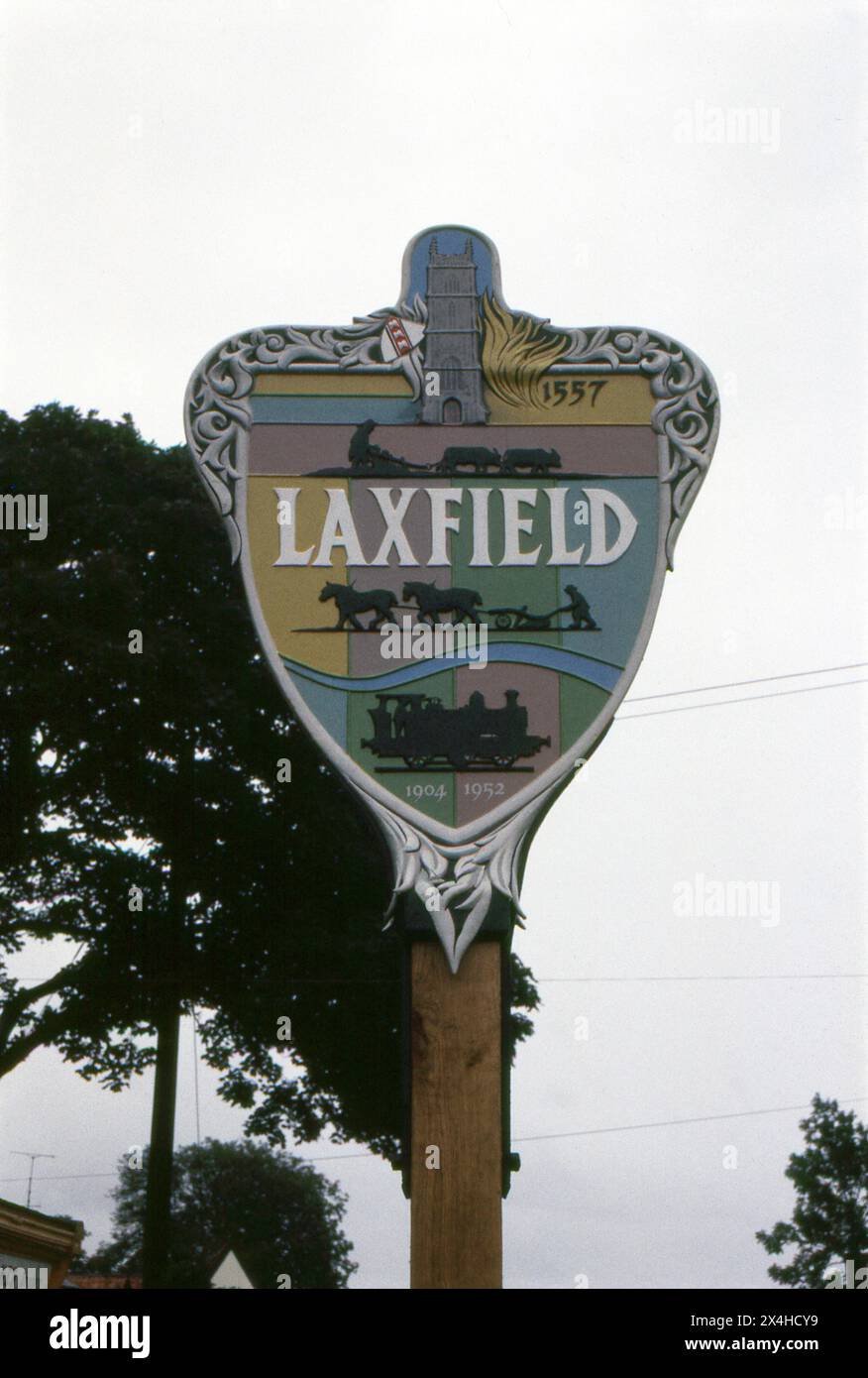 Suffolk. 1987: Ein Foto des Dorfzeichens Laxfield in Northern Suffolk, England. Das Schild ist aus geschnitztem Holz gefertigt und mit verschiedenen lokalen Wahrzeichen und Motiven verziert, darunter eine Dampflokomotive und Pflüger. Laxfield liegt an der B1117 zwischen Ashfield Green und Ubbeston Green. Stockfoto