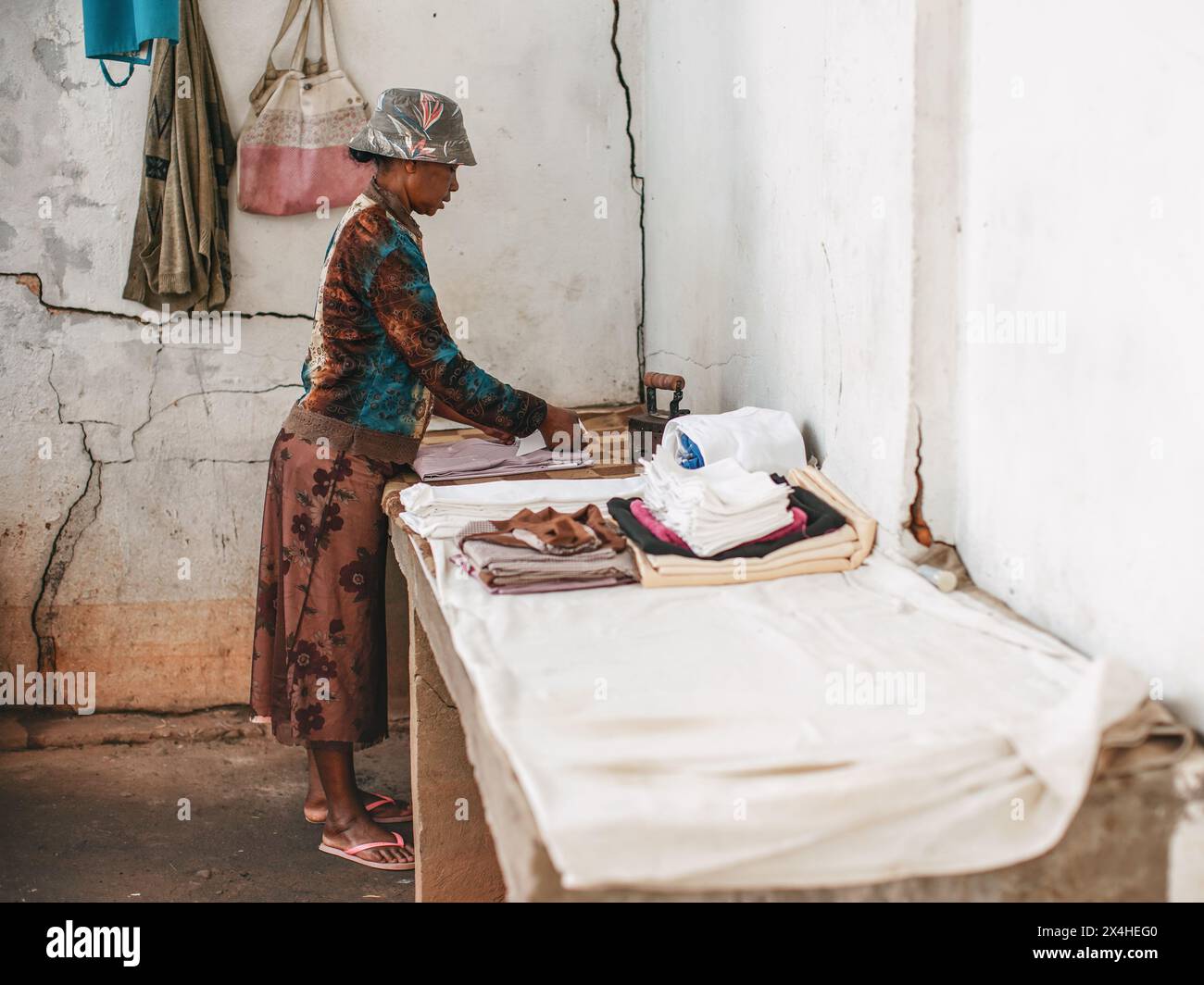 Ambositra, Madagaskar - 26. April 2019: Unbekannte lokale madagassische Frau, Bügelwäsche mit einfachem Bügeleisen, gebrochener weißer Hauswand Hintergrund Stockfoto