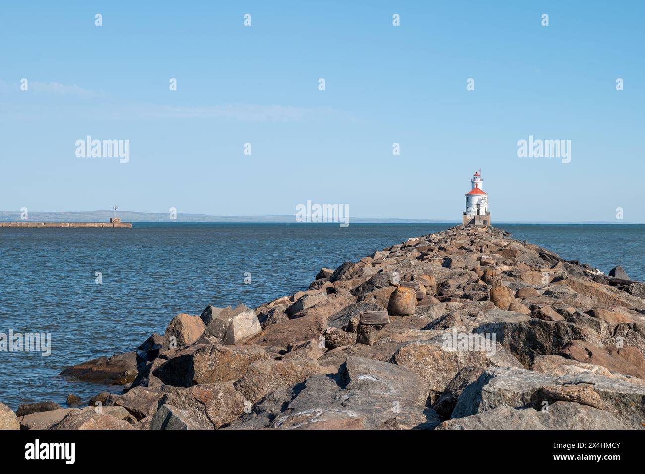 Leuchtturm mit rotem Dach am Ende eines felsigen Piers am Lake Superior, mit ruhigem Wasser und klarem blauem Himmel. Ein weiterer Pier ragt auf dem ins Wasser Stockfoto