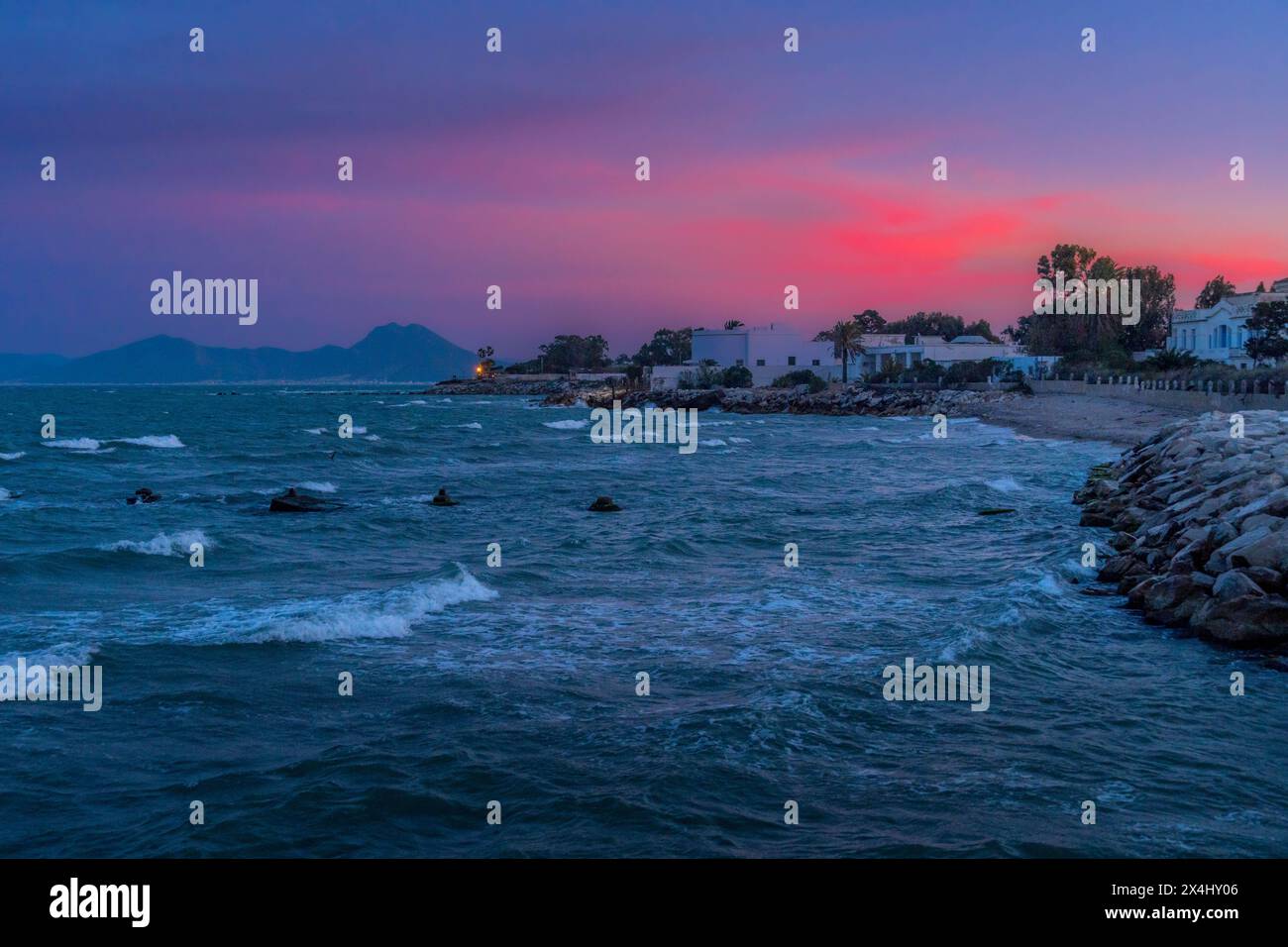 Der farbenfrohe Sonnenuntergang an Tunesiens Karthago Uferpromenade mit den winzigen Wolken, Wellen und Bergen am Horizont. Stockfoto