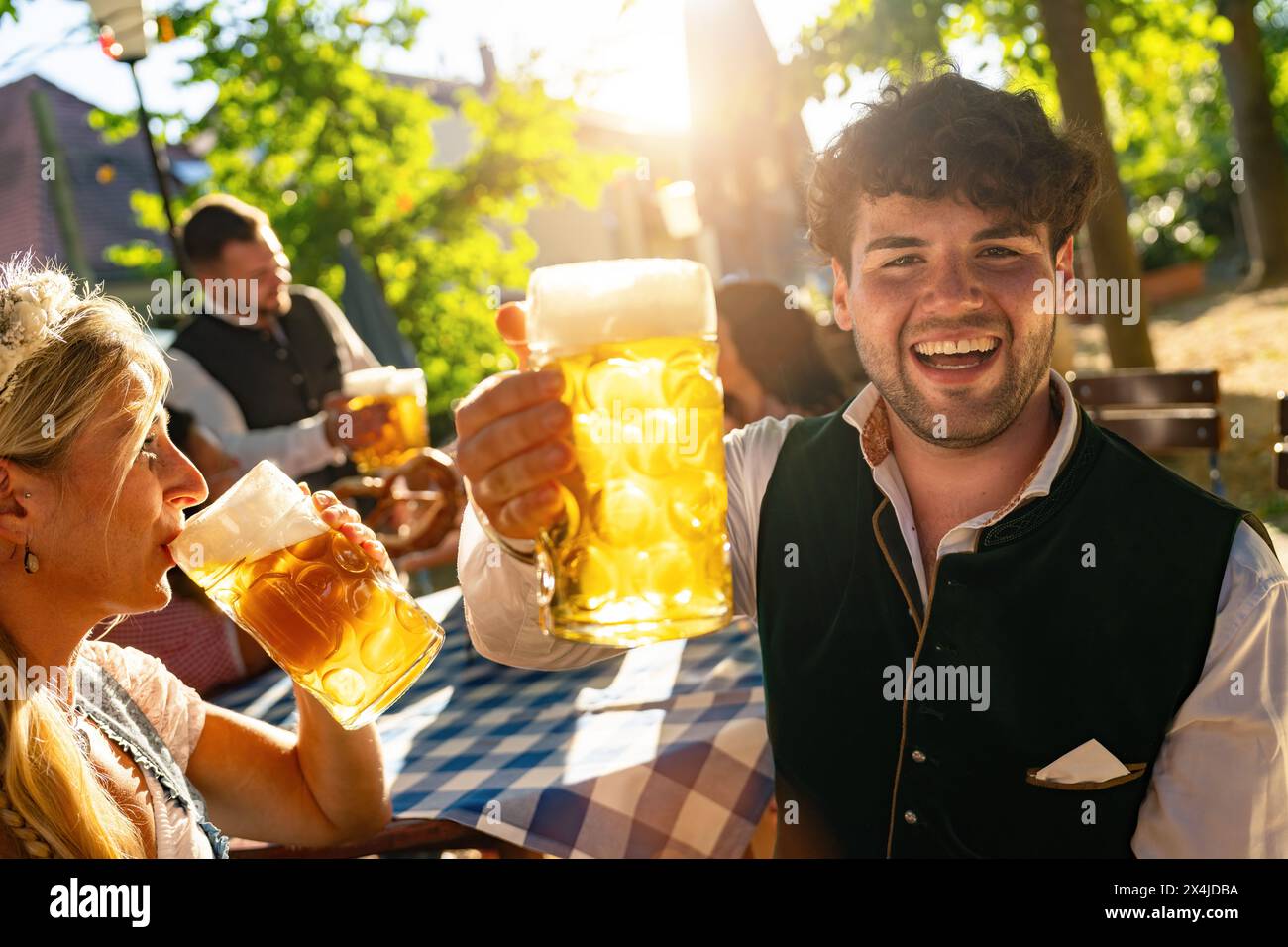 Fröhlicher junger Mann, der auf dem Oktoberfest oder im Biergarten mit einem Bierbecher tobt, in traditioneller Kleidung oder tracht auf Deutsch gekleidet, helles Sonnenlicht im Hintergrund Stockfoto