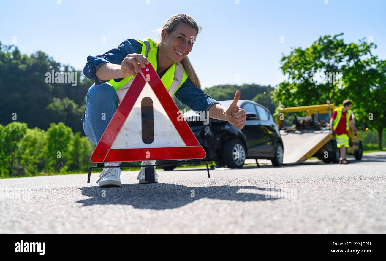 Frau, die ein Warndreieck auf der Straße aufbaut und ist glücklich und zufrieden mit einem schnellen und zuverlässigen Abschleppdienst für Hilfe auf der Straße. Sie zeigt do Stockfoto
