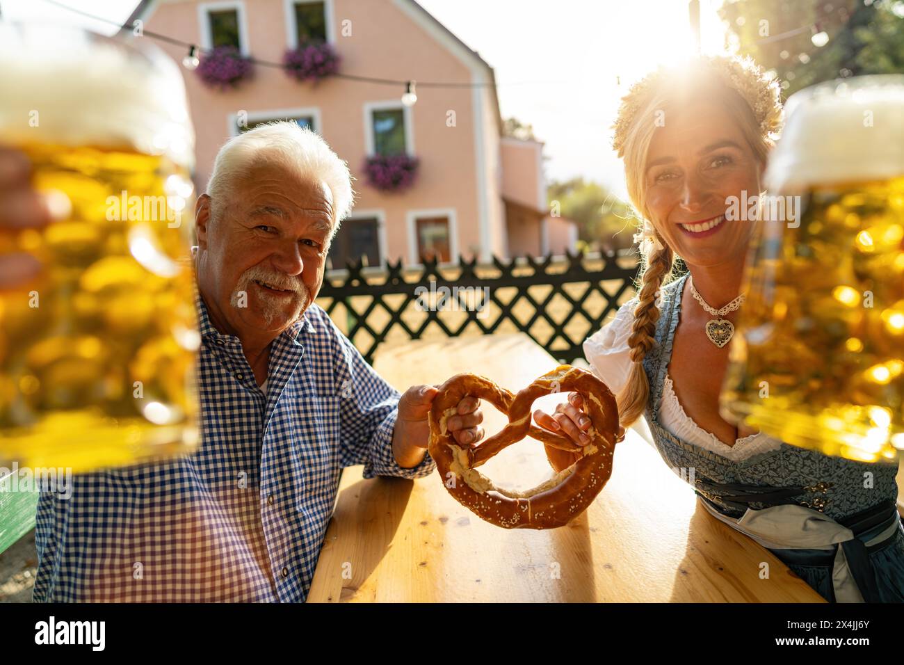 Seniorenmann und junge Frau in bayerischer Kleidung, die Bierbecher und Brezel halten, lächeln in einem Biergarten oder Oktoberfest in München Stockfoto