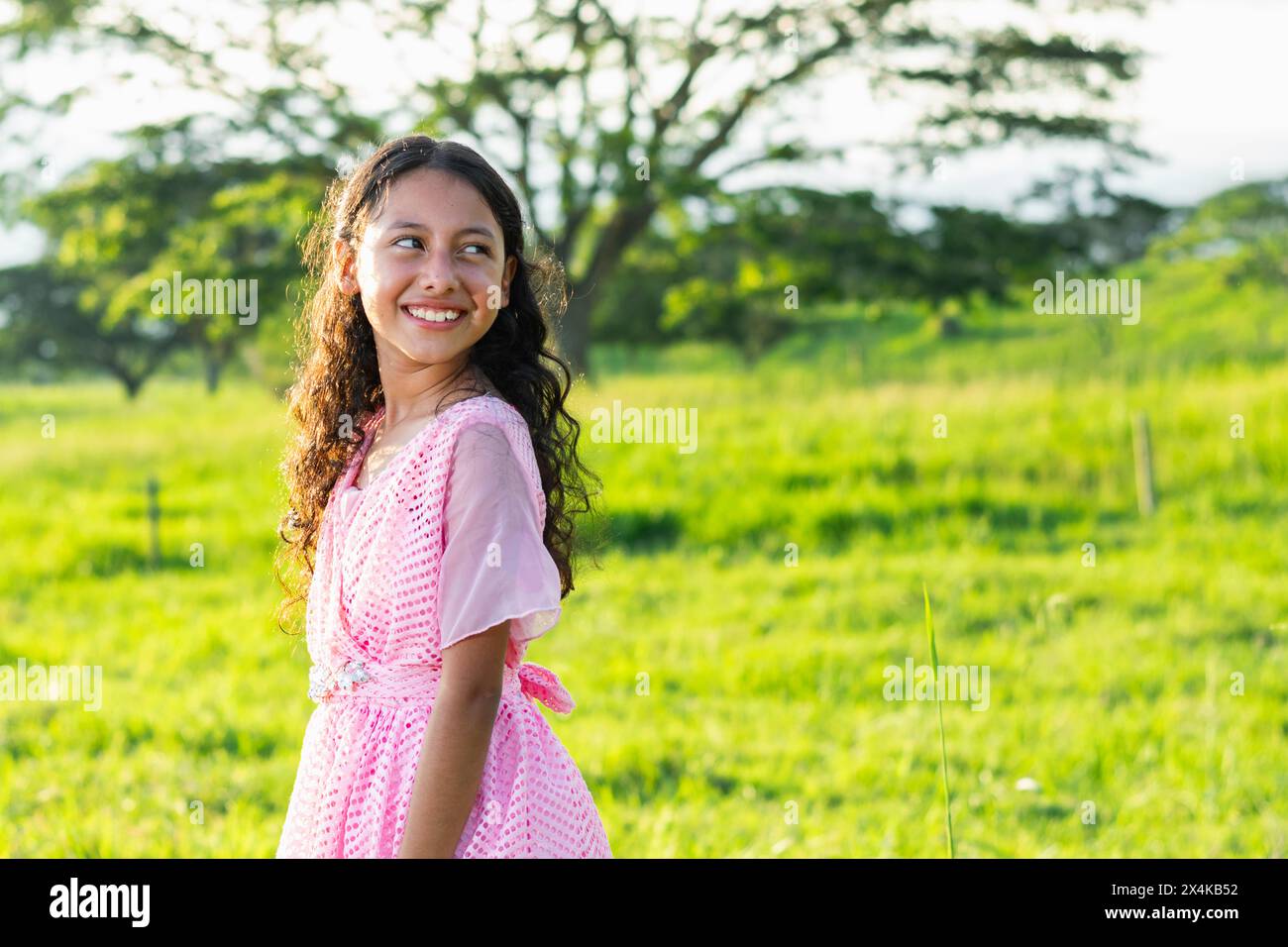 Das kleine brünette Latina Mädchen, das durch ein grünes Grasfeld geht, blickt zurück und lächelt vor großem Glück. Stockfoto