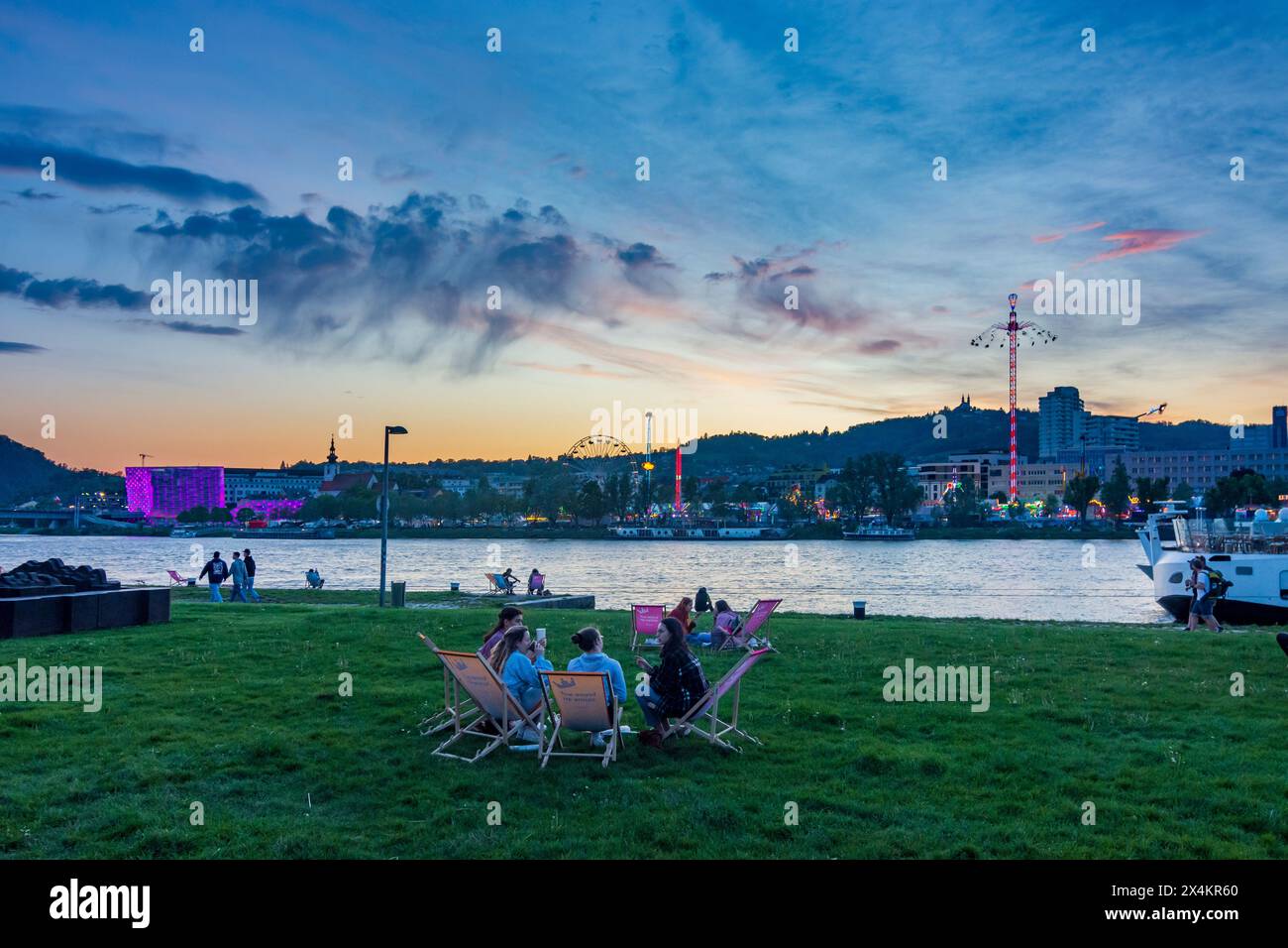 Linz: Donau (Donau), Kreuzfahrtschiff, Menschen sitzen auf der Wiese, Festival Urfahranermarkt mit Tower Swinger Star Flyer im Stadtteil Urfahr, Sonnenuntergang i Stockfoto