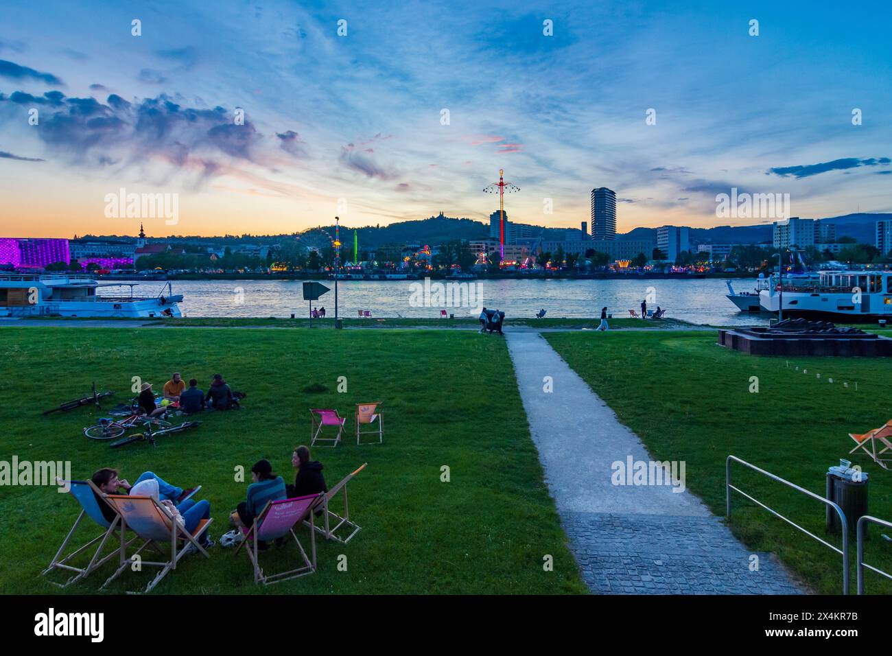 Linz: Donau (Donau), Kreuzfahrtschiff, Menschen sitzen auf der Wiese, Festival Urfahranermarkt mit Tower Swinger Star Flyer im Stadtteil Urfahr, Sonnenuntergang i Stockfoto