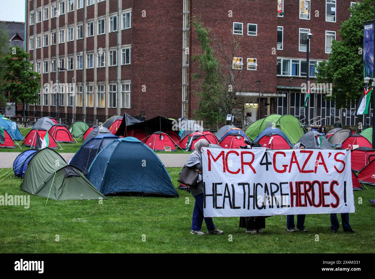 Die Demonstranten halten ein großes Banner mit der Aufschrift „MCR 4 Gaza's Healthcare Heroes“ während einer Mahnwache für die Beschäftigten des Gesundheitswesens in Palästina im Lager des Widerstands. Studenten und Unterstützer besetzen den Brunswick Park an der Manchester University. Die Demonstranten fordern, dass die Universität aufhört, Israel zu bewaffnen und ihre Mittäterschaft am Völkermord zu beenden. Sie bestehen darauf, dass die Universität ihre Partnerschaft mit dem Waffenhersteller BAE Systems beendet, ihre Beziehungen zu Tel Aviv und den Hebräischen Universitäten in Israel beendet und eine Politik verfolgt, die sicherstellt, dass alle Forschungsarbeiten ethisch sind und nicht zum Waffenhandel beitragen. Die Demonstranten sind entschlossen, hier zu bleiben Stockfoto