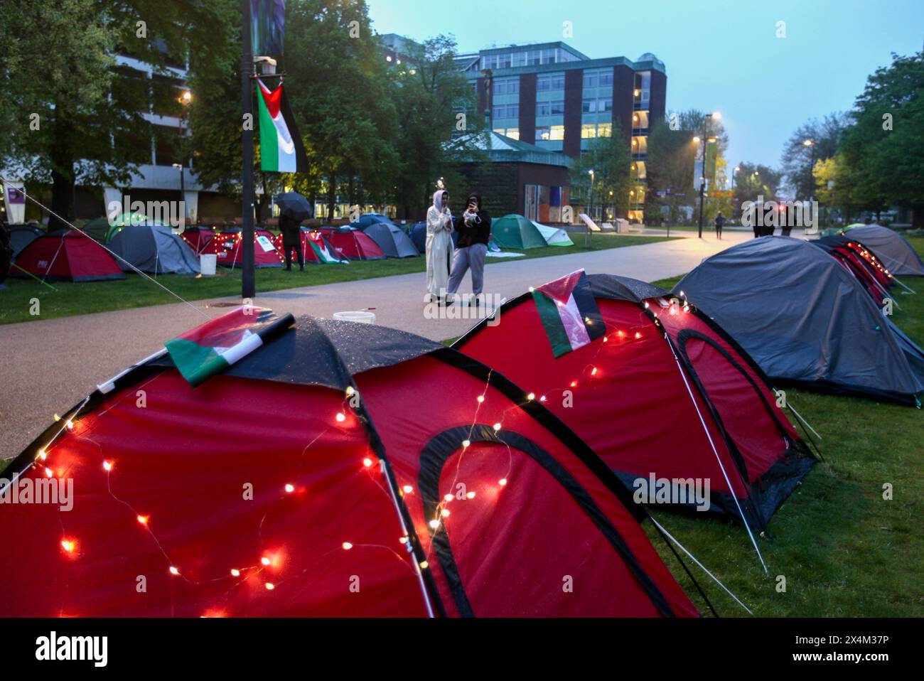 Feenlichter zieren einige der Zelte und eine palästinensische Flagge fliegt bei Einbruch der Dunkelheit über dem Lager des Widerstands für Palästina. Studenten und Unterstützer besetzen den Brunswick Park an der Manchester University. Die Demonstranten fordern, dass die Universität aufhört, Israel zu bewaffnen und ihre Mittäterschaft am Völkermord zu beenden. Sie bestehen darauf, dass die Universität ihre Partnerschaft mit dem Waffenhersteller BAE Systems beendet, ihre Beziehungen zu Tel Aviv und den Hebräischen Universitäten in Israel beendet und eine Politik verfolgt, die sicherstellt, dass alle Forschungsarbeiten ethisch sind und nicht zum Waffenhandel beitragen. Die Demonstranten sind entschlossen, bis zu ihrem Dekan in der Besatzung zu bleiben Stockfoto