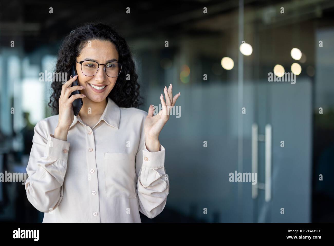Glückliche erfolgreiche hispanische Frau, die am Telefon spricht, Geschäftsfrau am Arbeitsplatz im Büro, Arbeiter mit lockigen Haaren und Brillen, selbstbewusste Chefin. Stockfoto