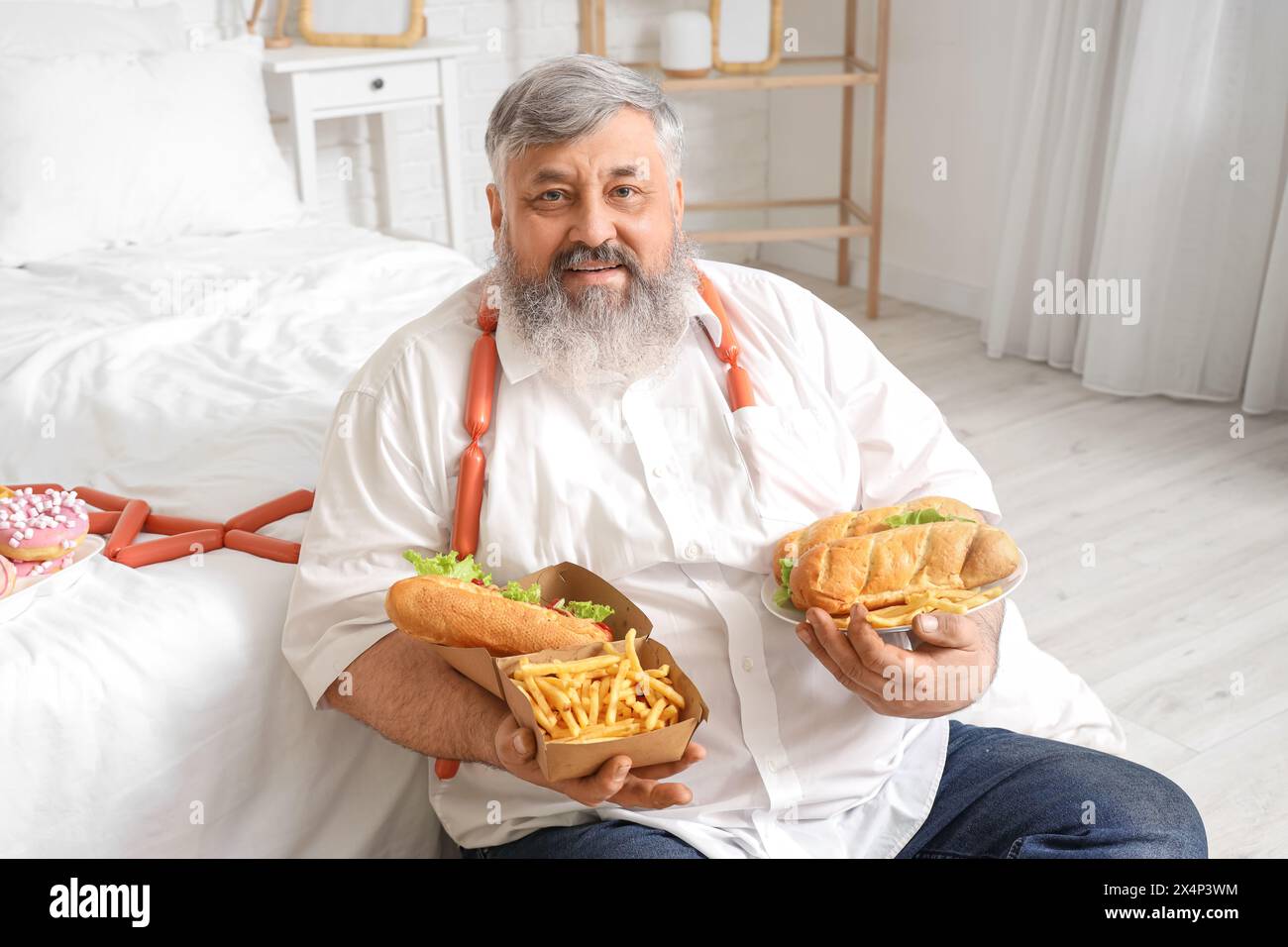 Übergewichtiger reifer Mann mit ungesunder Nahrung im Schlafzimmer. Das Konzept des Überessens Stockfoto