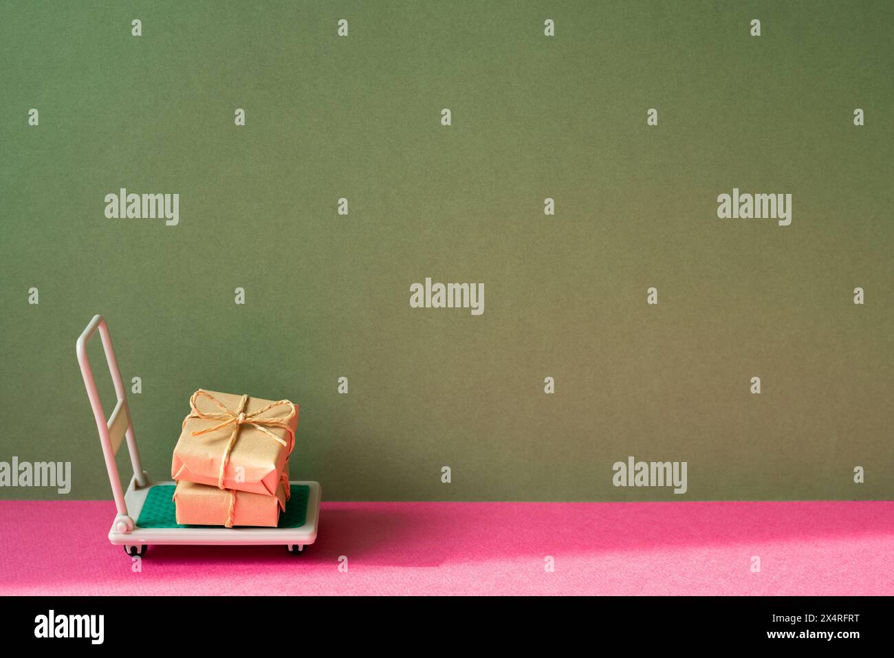 Trolley-Wagen mit Geschenkboxen auf rosa und khakigrünem Hintergrund. Paketzustellungskonzept Stockfoto