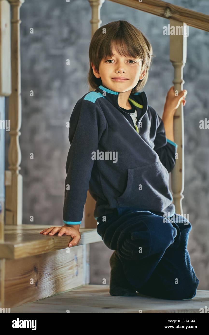 Der kaukasische Vorschuljunge von sieben Jahren posiert auf Stufen einer Holztreppe und hält Baluster mit der Hand. Stockfoto