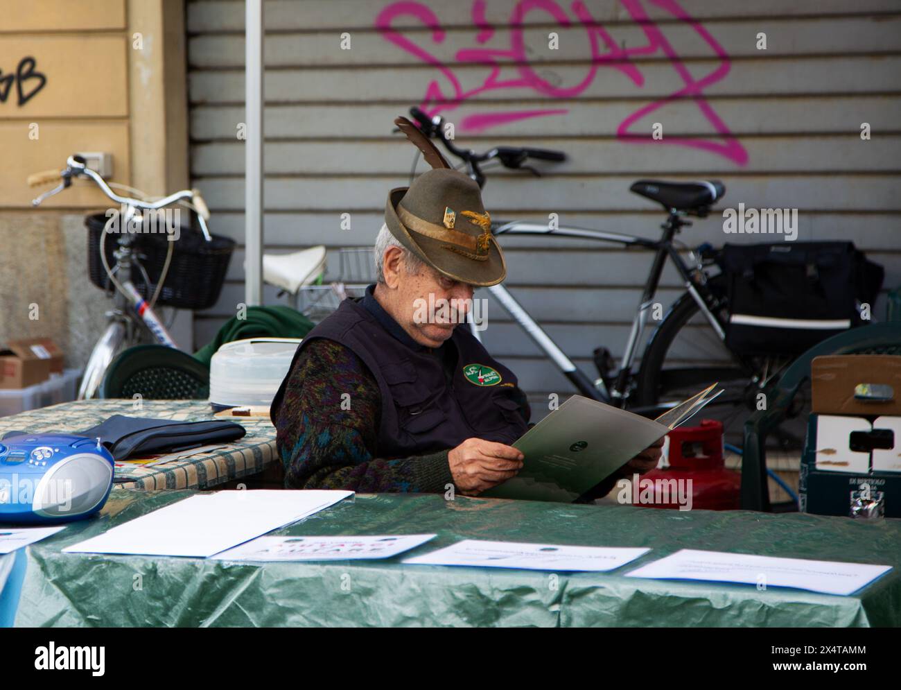 Ein Mann mit italienischem Alpenmilitärhut, der hinter der Theke beim Dorffest sitzt Stockfoto