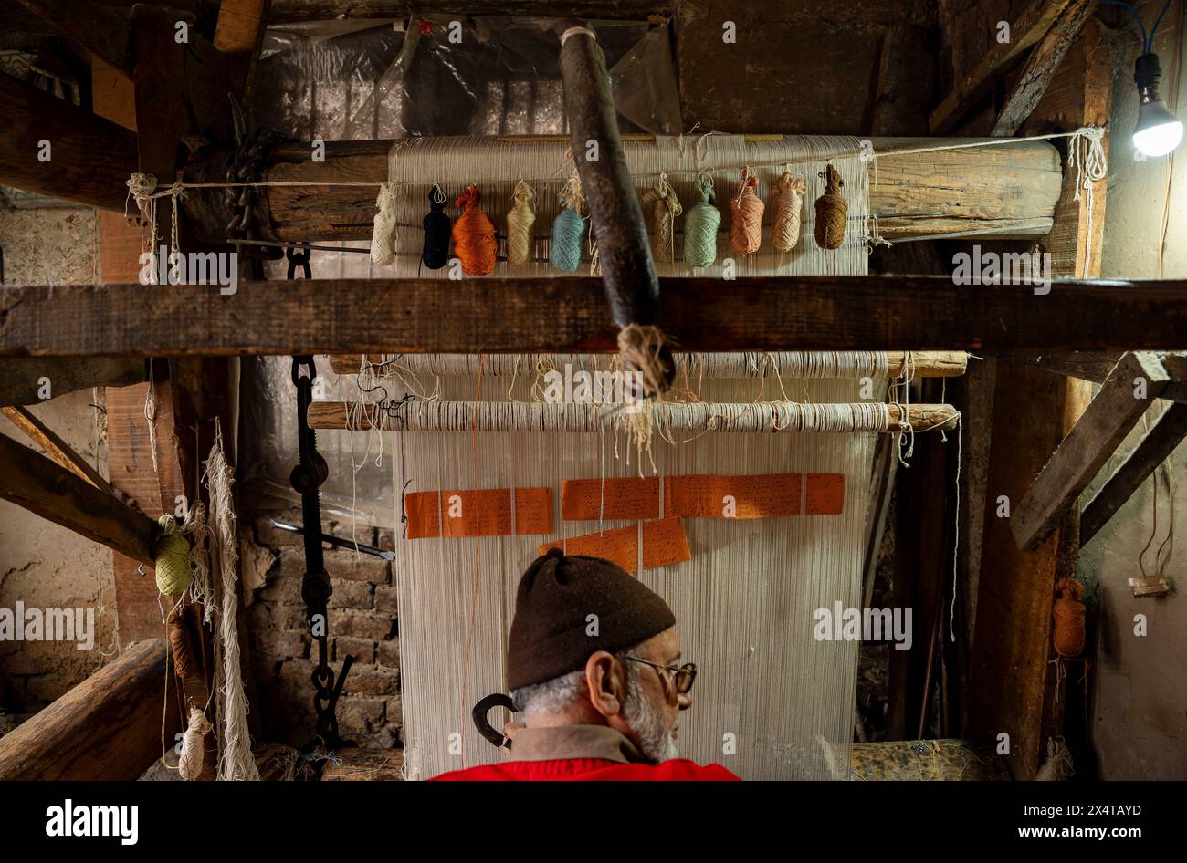 Neben einem Handwerker, der an einem traditionellen Teppich arbeitet, der in einer Teppichweberei gewebt wird, werden Entwürfe geschrieben. Die drei-Wege-Teppichfabrik in Srinagar, einst ein blühendes Zentrum der Handwerkskunst, ist heute ein Beweis für die Herausforderungen, denen die traditionellen Industrien in einer sich wandelnden Welt gegenüberstehen. Nur noch eine Handvoll Handwerker, darunter ein älterer Staatsmann, der die verblasste Tradition verkörpert, sind in den Hallen der Fabrik Erinnerungen an eine vergangene Ära zu finden. Die ehrlichen Reflexionen der älteren Handwerker zeigen die harten Realitäten des Niedergangs der Industrie: Das mangelnde Interesse der jüngeren Generation Stockfoto