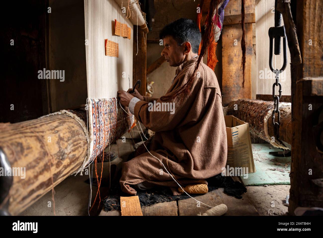 In einer Teppichweberei wird ein Kunsthandwerker aus Kaschmir gesehen, der einen traditionellen Teppich webt. Die drei-Wege-Teppichfabrik in Srinagar, einst ein blühendes Zentrum der Handwerkskunst, ist heute ein Beweis für die Herausforderungen, denen die traditionellen Industrien in einer sich wandelnden Welt gegenüberstehen. Nur noch eine Handvoll Handwerker, darunter ein älterer Staatsmann, der die verblasste Tradition verkörpert, sind in den Hallen der Fabrik Erinnerungen an eine vergangene Ära zu finden. Die ehrlichen Reflexionen der älteren Handwerker zeigen die harten Realitäten des Niedergangs der Branche: Mangelndes Interesse bei der jüngeren Generation, saisonale Schwankungen i Stockfoto