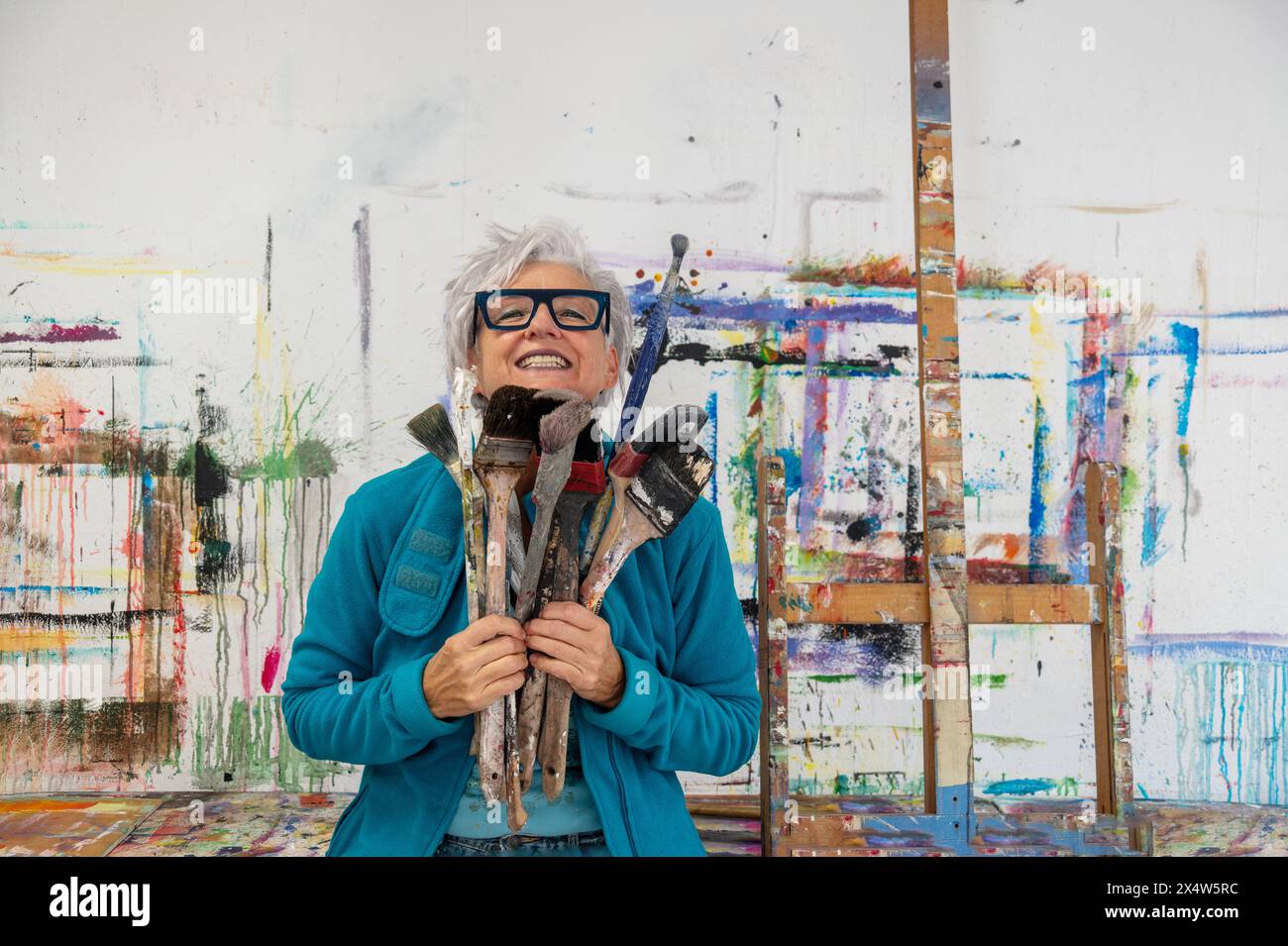 Ältere grauhaarige reife glückliche lächelnde Künstlerin stolze Künstlerin, in ihren fünfziger Jahren mit grauen Haaren und Brillen und großen Pinseln, in ihrem Atelier, Kopie Stockfoto