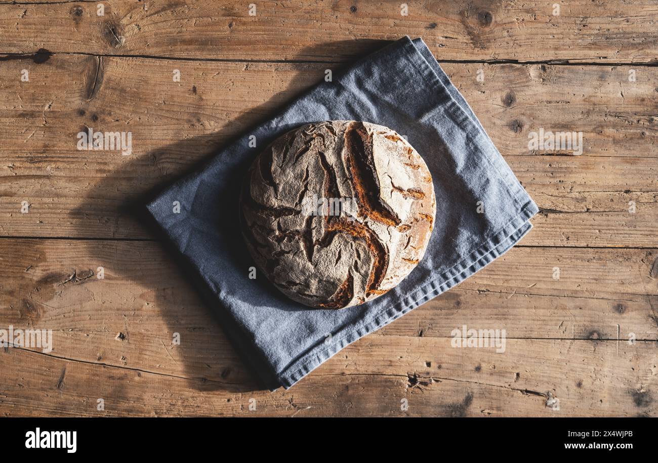 Frisch gebackener runder Brotlaib auf einem blauen Tuch, auf einem rustikalen Holztisch Stockfoto