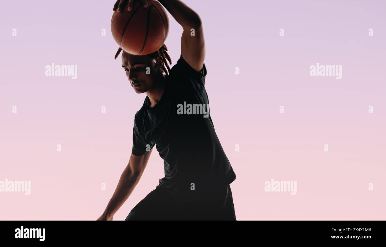 Ein junger Athlet mit Dreadlocks dribbelt einen Basketball in einem Studio. Seine Form ist auf den Punkt gekommen, während er beleidigt, zeigt Geschick und Leichtathletik. Stockfoto