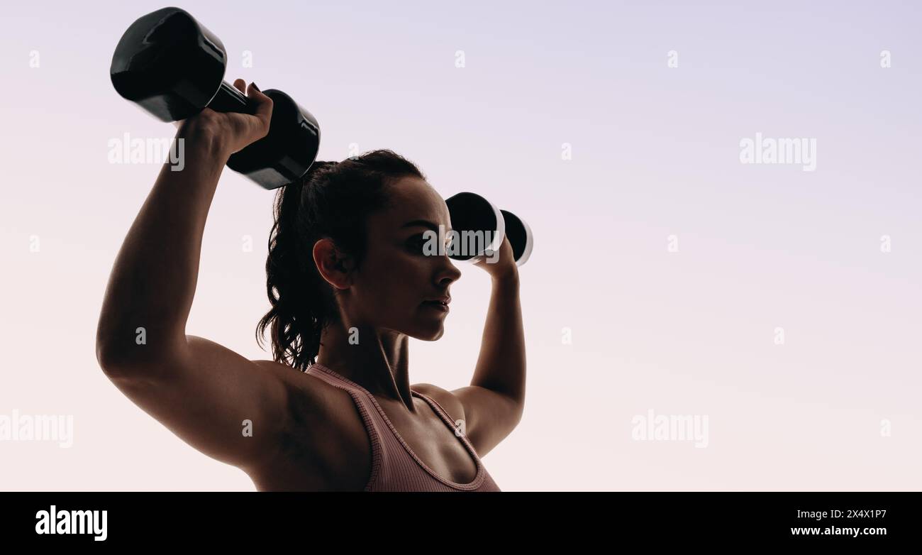 Eine fitte Frau in Sportbekleidung trainiert ihren Oberkörper in einem Studio, hebt Gewichte und benutzt Kurzhanteln. Ihre Silhouette zeigt ihren straffen Körper. Stockfoto