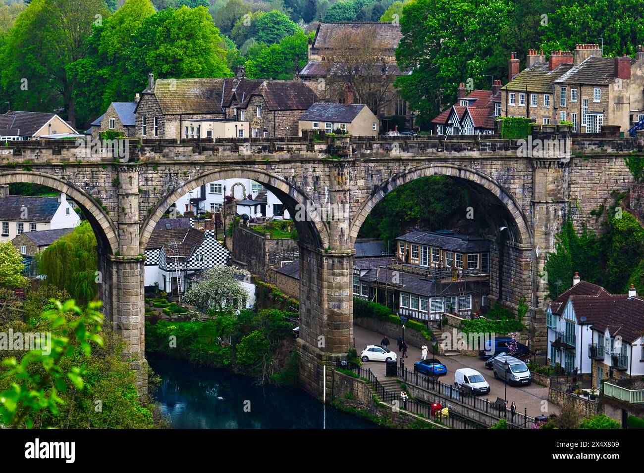 Ein malerischer Blick auf eine Steinbogenbrücke über einen Fluss, umgeben von üppigem Grün und malerischen Häusern in einem charmanten Dorf. Stockfoto