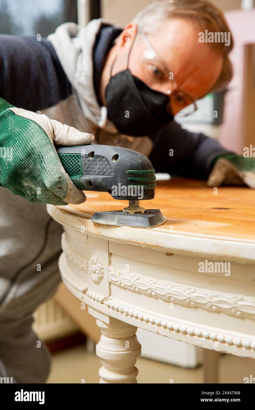 Ein Mann mittleren Alters, der alten Holztisch mit einem elektrischen Schleifgerät hochfährt. Möbelsanierung, Heimwerkerreparatur, Eigenreparatur und Wohnungsverbesserung. Recycling Stockfoto