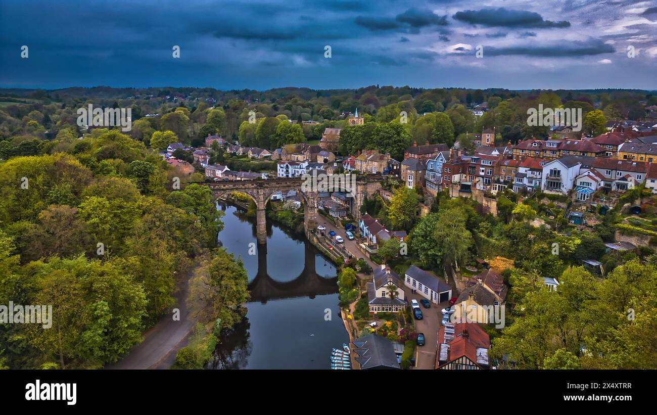 Aus der Vogelperspektive auf eine malerische Stadt mit einer historischen Steinbrücke über einen Fluss, umgeben von üppigem Grün und Wohnhäusern unter einem dramatischen Himmel. Stockfoto