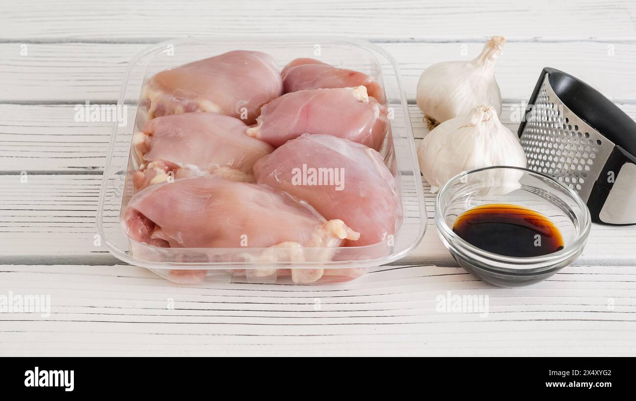 Rohe Hähnchenschenkel in einem Plastikbehälter, Sojasauce und frischer Knoblauch und Knoblauch drücken Nahaufnahme auf einem weißen Holzhintergrund Stockfoto