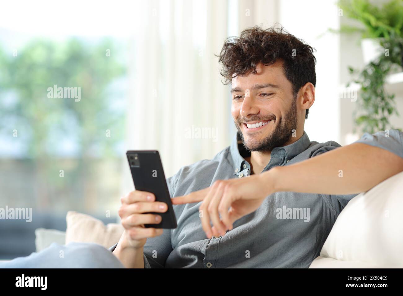 Glücklicher Mann, der sein Handy benutzt und zu Hause auf einer Couch sitzt Stockfoto