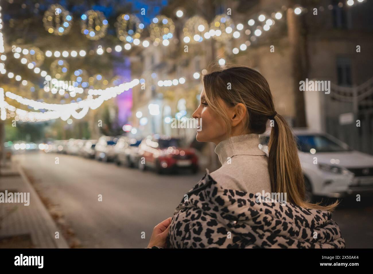 Eine Frau mit Leopardenmotiv steht nachts auf der Straße. Die Straße ist mit Weihnachtslichtern beleuchtet, was eine festliche Atmosphäre schafft. Es gibt Stockfoto