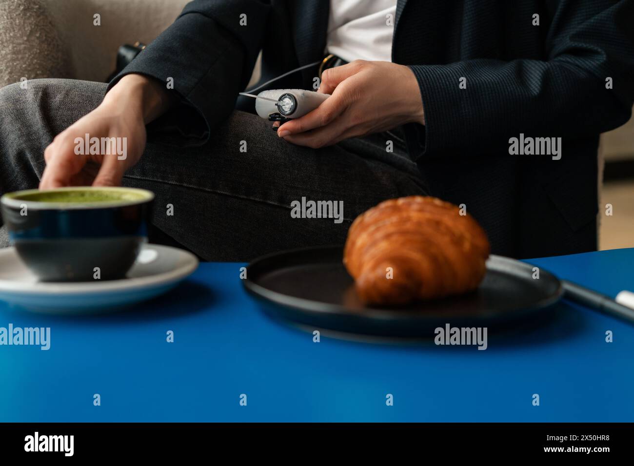 Nahaufnahme einer Frau, die den Blutzuckerspiegel in einem Café überprüft, bevor sie ein Croissant isst und einen Matcha-Tee trinkt Stockfoto