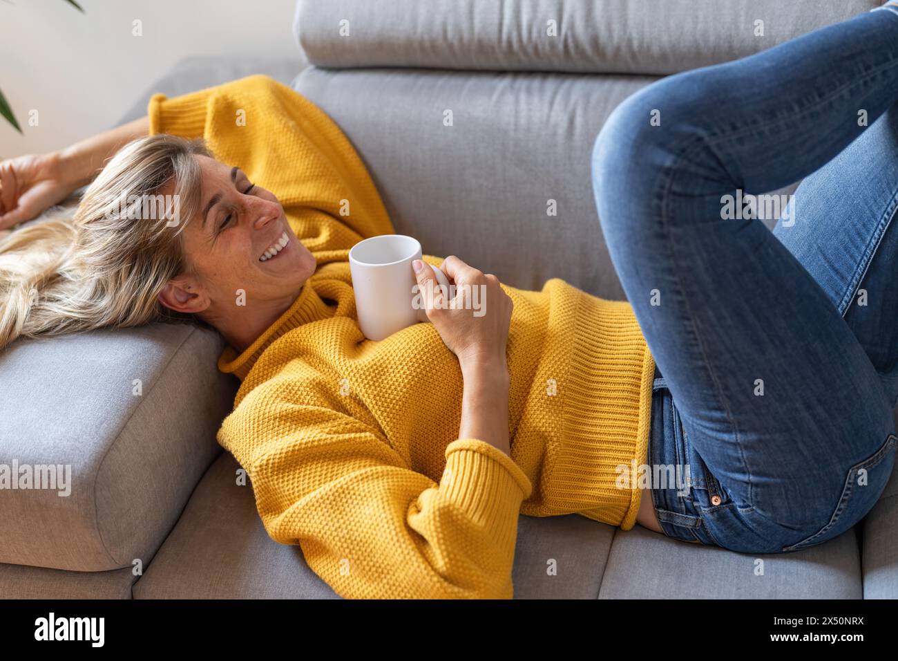 Reife Frau, die sich zu Hause auf der Couch mit einer Kaffeetasse entspannt, lächelnd und fröhlich aussah Stockfoto