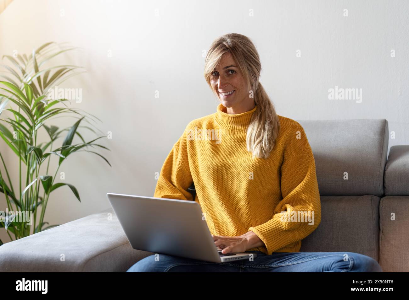 Lächelnde junge Frau, die auf einem Sofa sitzt und einen Laptop benutzt, mit einer Zimmerpflanze neben ihr Stockfoto