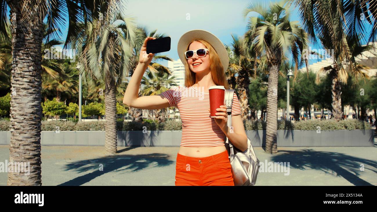 Porträt einer jungen Frau, die Selfie mit Telefon im Sommerpark macht, mit Rucksack, Strohhut vor Palmen Hintergrund Stockfoto