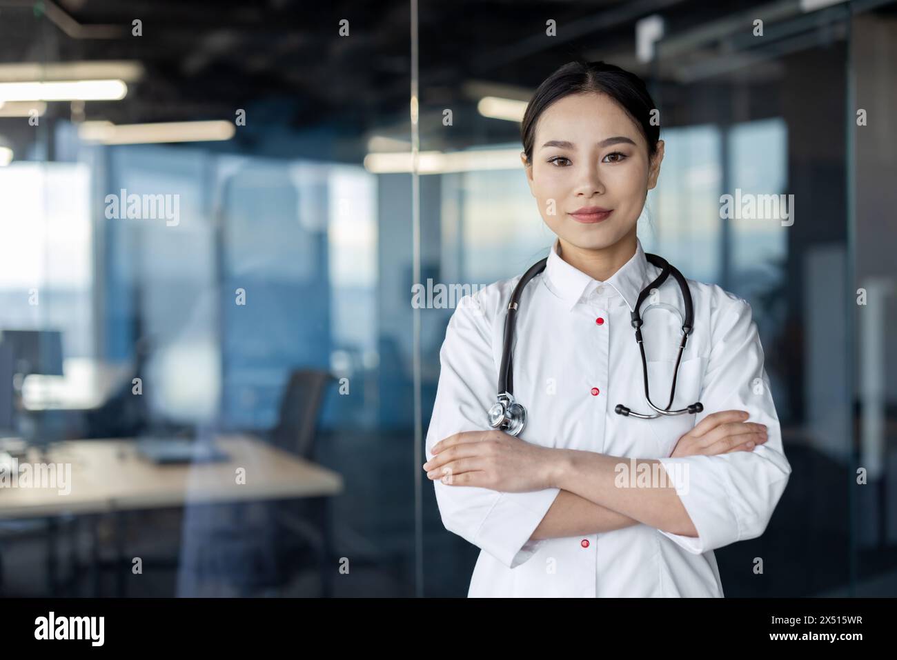 Eine ernsthafte asiatische Ärztin mit gekreuzten Armen, die selbstbewusst in einer modernen Krankenhausumgebung steht und Professionalität und Bereitschaft unter Beweis stellt. Stockfoto