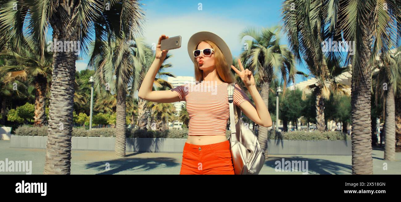 Porträt einer jungen Frau, die Selfie mit Telefon im Sommerpark macht, mit Rucksack, Strohhut vor Palmen Hintergrund Stockfoto