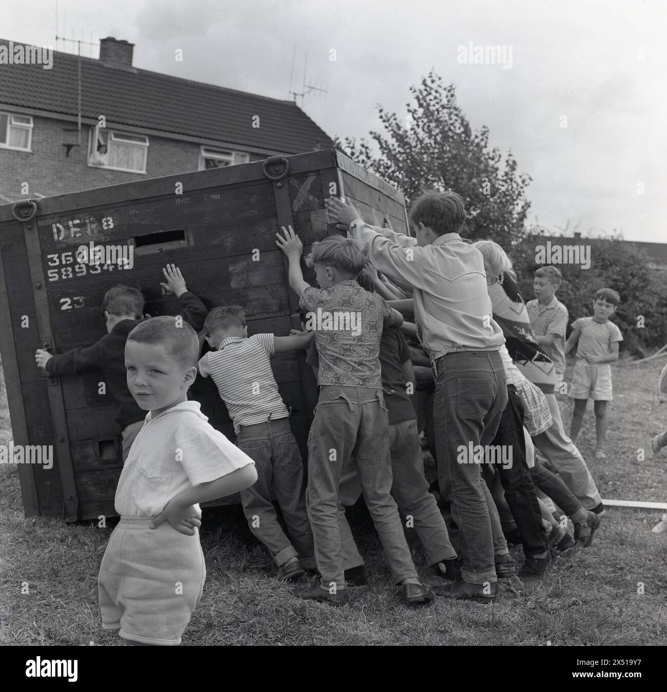 1964, historisch, draußen auf einem Feld, eine Gruppe von Kindern, die eine große Holz-/Metall-Fracht-Kiste auf die Seite schieben, um zu spielen und Spaß zu haben. Stockfoto