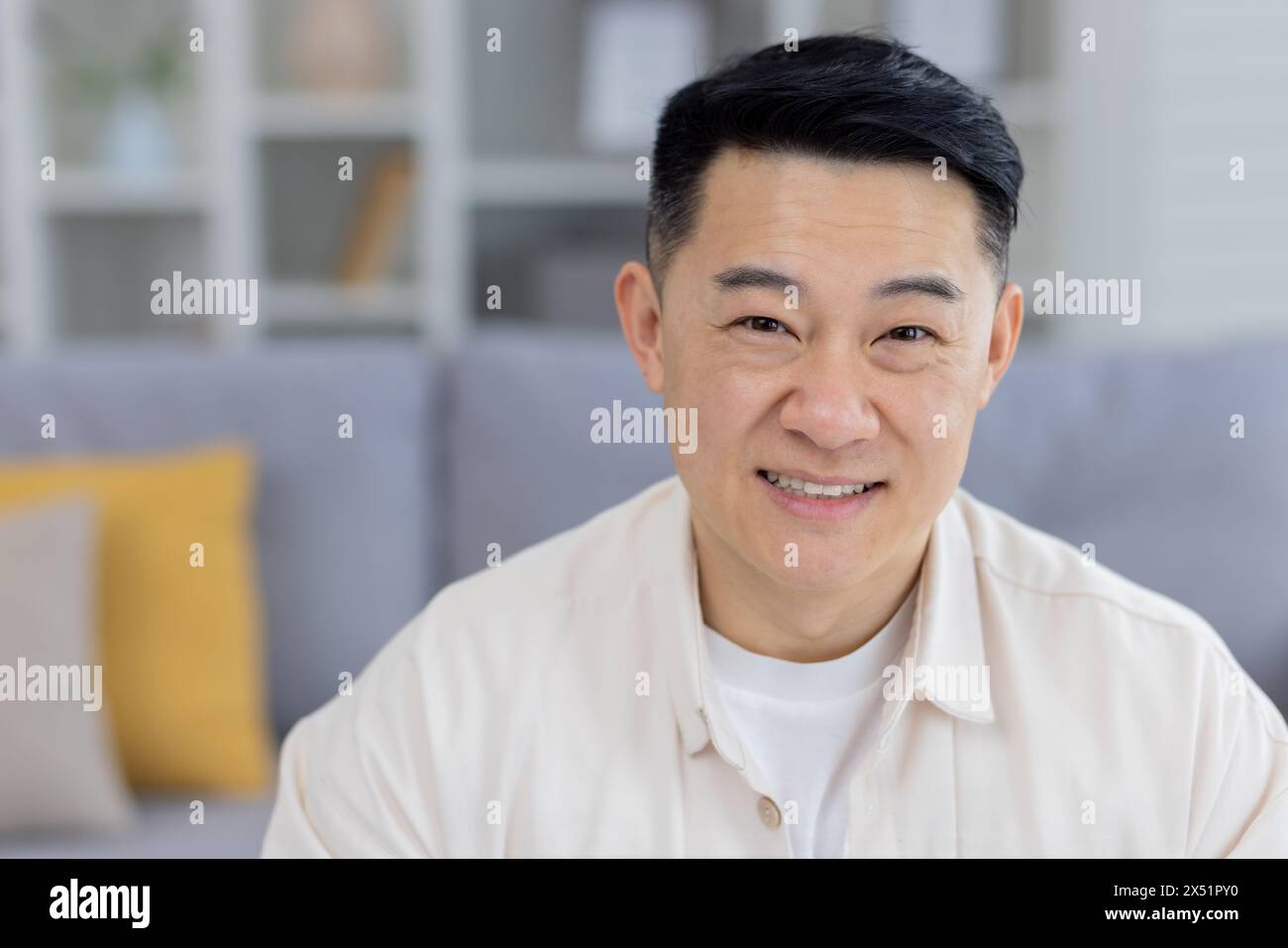 Porträt eines glücklichen asiatischen Mannes mittleren Alters, der in einem modernen Wohnzimmer sitzt und ein lässiges cremefarbenes Hemd mit einem entspannten, selbstbewussten Lächeln trägt. Stockfoto