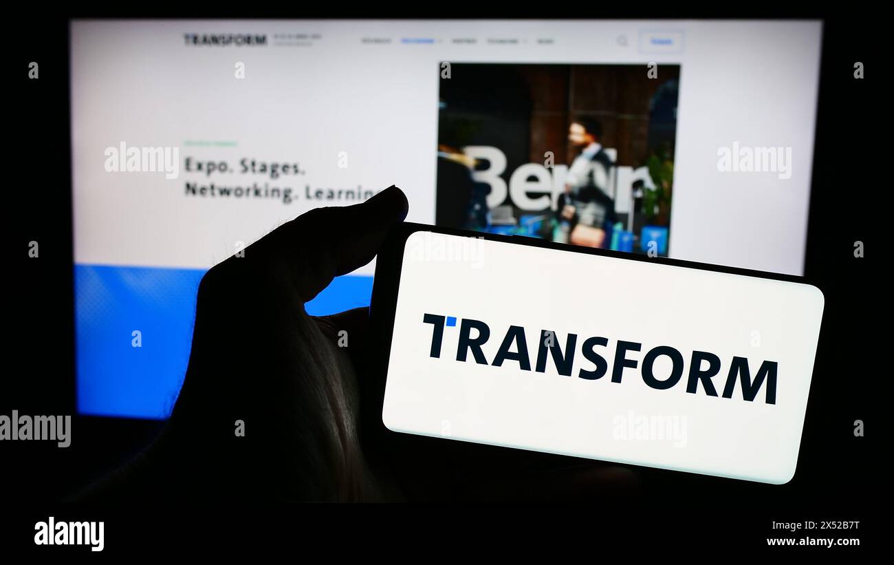 Person mit Smartphone mit Logo des deutschen Technologieereignisses TRANSFORMATION (Bitkom e.V.) vor der Website. Schwerpunkt auf dem Display des Telefons. Stockfoto