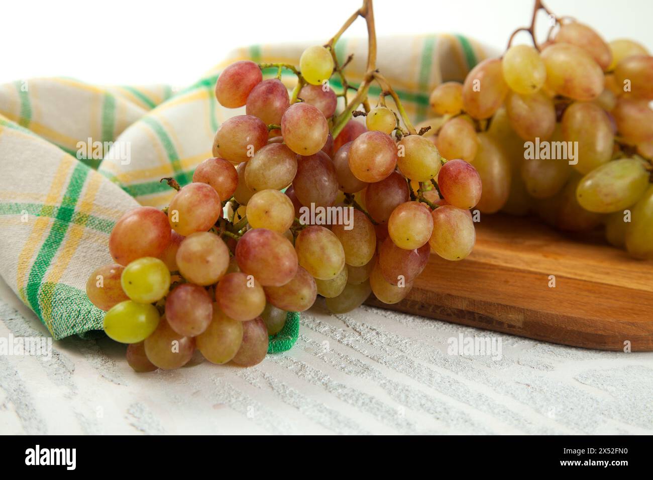Schneidebrett mit einem großen Haufen reifer rosafarbener Trauben auf weiß. Rosafarbene Weintrauben mit blauem Küchentuch auf weißem hölzernem Hintergrund. Stockfoto