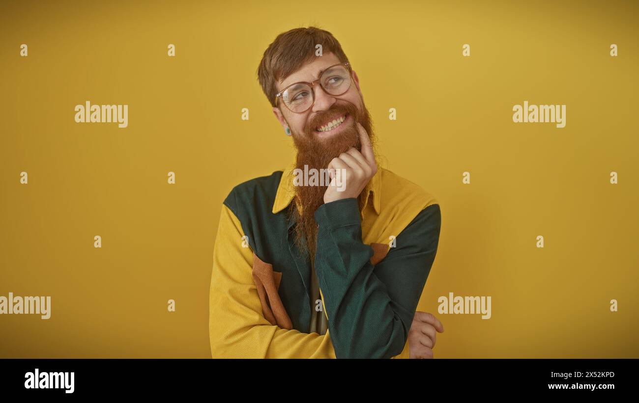Ein lächelnder bärtiger rothaariger Mann in Freizeitkleidung posiert nachdenklich vor gelbem Hintergrund. Stockfoto