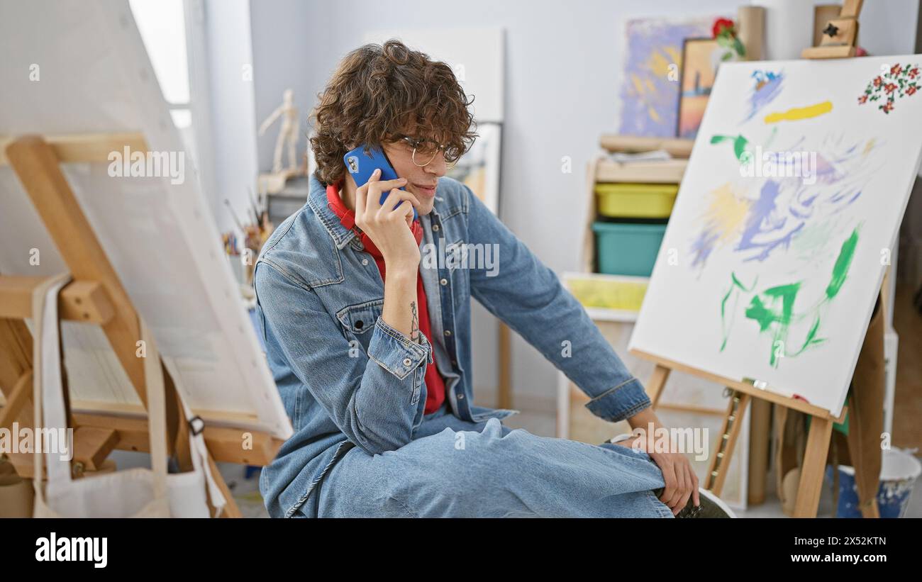 Ein stilvoller Mann mit lockigen Haaren spricht am Telefon in einem Kunststudio, umgeben von Leinwänden und Gemälden. Stockfoto