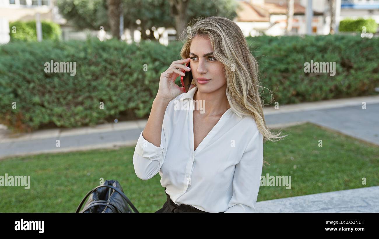 Coole, lässige und konzentrierte, blonde junge Frau im sonnigen Stadtpark mit ihrem Smartphone Stockfoto