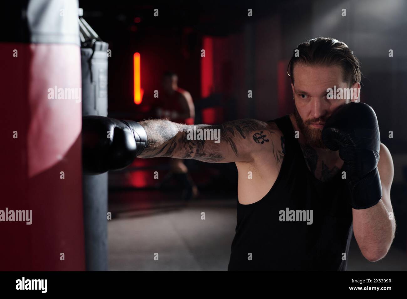 Bärtiger muskulöser Mann mit Tätowierungen auf Arm und Brust, der vor dem Boxsack steht und ihn während des Trainings im Fitnessstudio oder Boxring trifft Stockfoto