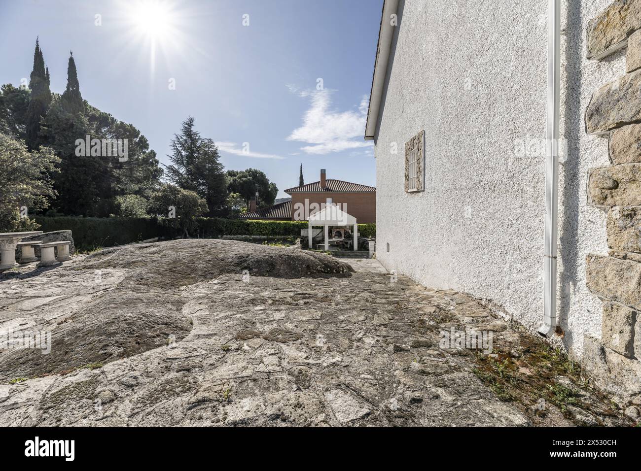 Einfamilienhaus mit einer weißen und Steinfassade in der Mitte eines landschaftlich gestalteten Anwesens mit Umzäunung und großen Flächen mit grauem Granit gepflastert Stockfoto