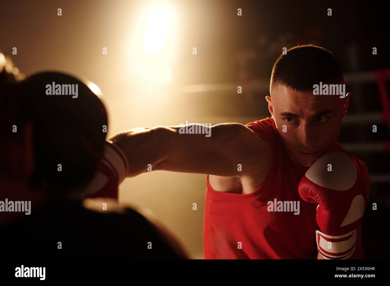 Junger muskulöser Mann in roter Sportbekleidung und Boxhandschuhen, der seinen Rivalen ansieht, während er ihn während der Runde am Ring auf den Kopf tritt Stockfoto