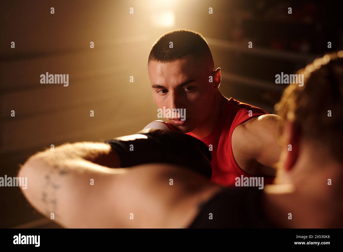 Junger männlicher Boxer, der Rivalen während des Kampfes oder der Verteidigung auf dem Boxring mit Licht und Rauch auf dem Hintergrund schlägt Stockfoto