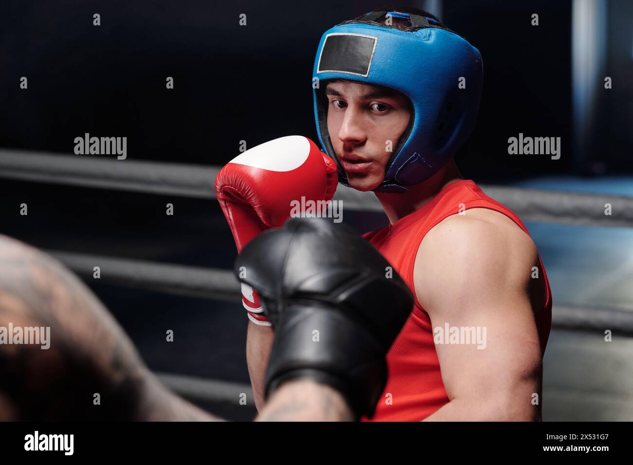 Junger muskulöser Mann in roter Weste und Boxhandschuhen und blauem Gesichtsschild, der seinen Rivalen ansieht, bevor er während des Kampfes auf dem Ring tritt Stockfoto