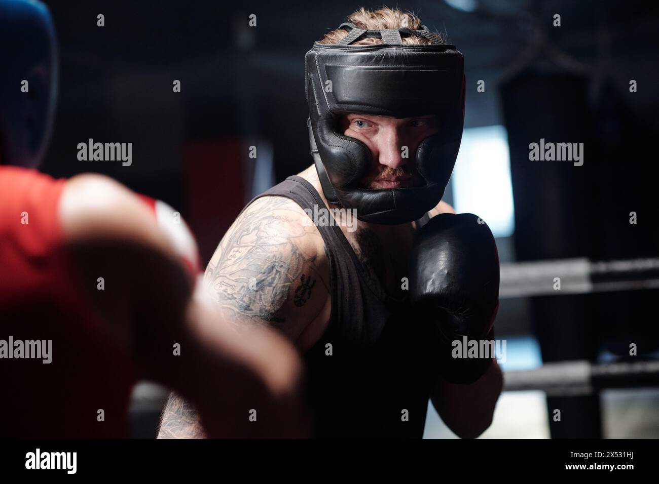 Junger männlicher Boxer in schwarzer Weste, Gesichtsschutz und Boxhandschuhen, der vor seinem Rivalen auf einem Ring steht und ihn zwingt Stockfoto