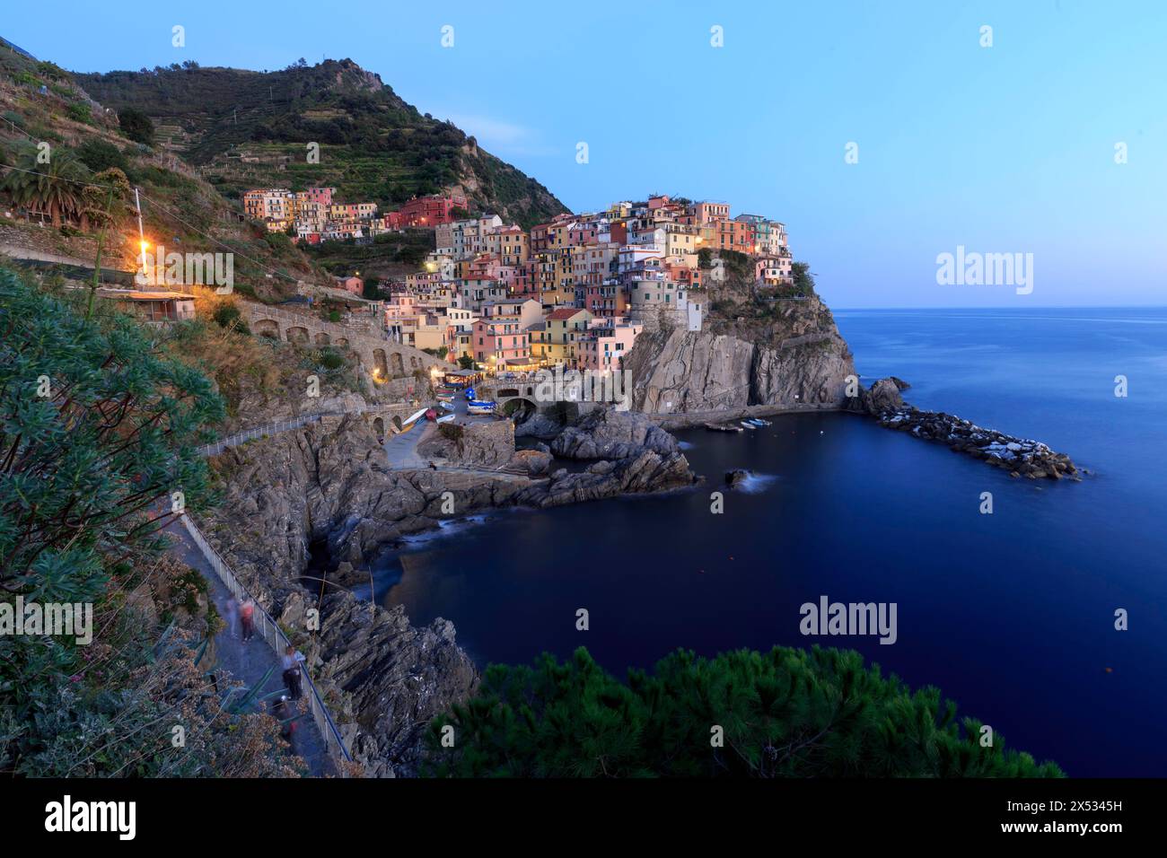 Mediterranes Dorf auf Felsen an der Küste in der Abenddämmerung mit eingeschalteten Lichtern und ruhigem Meer, Italien, Ligurien, Manarola, Riomaggiore, Provinz La Spezia Stockfoto